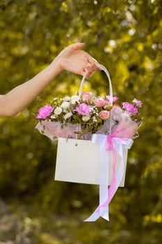 A beautiful bouquet of flowers in a box in the hands of a beautiful girl who walks along the street on a sunny day against the backdrop of green plants. Focus on the background of flowers.