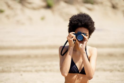 African American female photographer in swimwear taking picture on photo camera while standing on sandy shore during trip in tropical resort