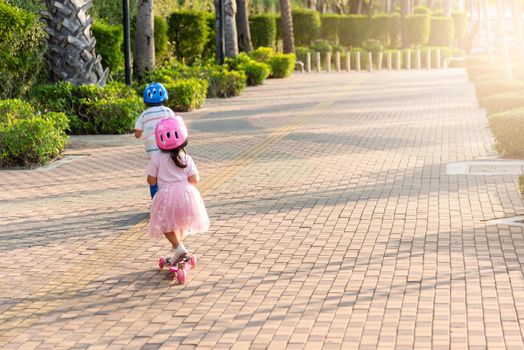 Child riding scooter. Back Asian little kid boy and girl wear safe helmet play kick board on road in park outdoors on summer day, Active children games outside, Kids sport healthy lifestyle concept