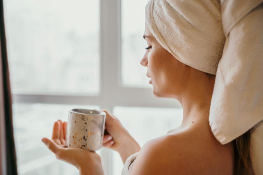 Young serene relaxed woman in spa bath towel drinking hot beverage tea coffee after taking shower bath at home. Beauty treatment, hydration concept