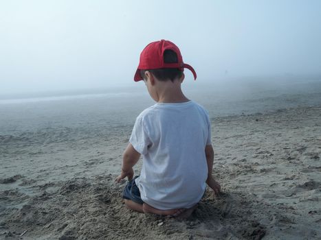 Boy sitting at the beach with his back to camera. Foggy sea view.