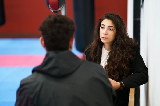 Young brunette with long wavy hair sitting on bench and listening to male athlete during therapy session in gym