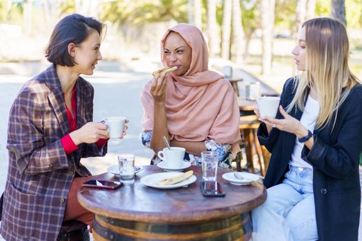 Group of multiracial female friends having lunch together at table with cups of coffee on terrace of outdoor cafe in city