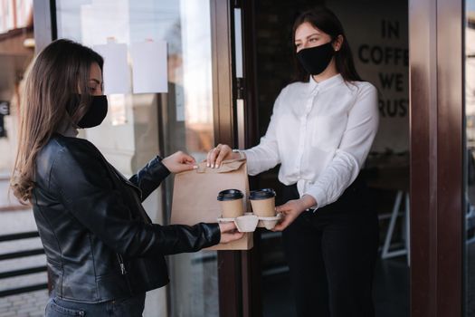 Young woman standing outdoors in mask and paying bill through smartphone using NFC technology in a cafe. Female customer paying using contactless technology. Woman taking at order. Take away food concept.