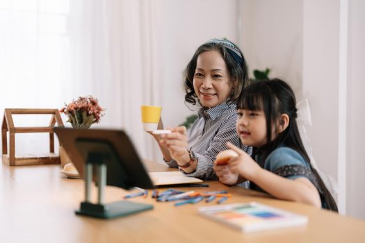 Smiling senior grandma and cute little kid granddaughter watching cartoons on laptop together, happy older grandmother grawing and painting