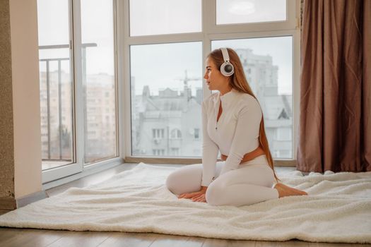 Side view portrait of relaxed woman listening to music with headphones lying on carpet at home. She is dressed in a white tracksuit