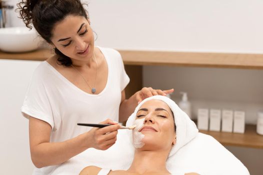 Smiling female cosmetologist applying hydrating mask with brush on face of female customer lying on table in beauty salon