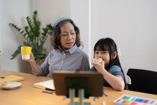 Mature grandmother helping to granddaughter with school assignment, focused little girl writing notes, older teacher training pupil, involved in lesson at home, homeschooling concept.