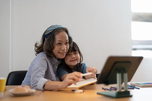 Loving senior grandmother hug shoulders of small girl grandchild assist in preparing homework support give advice. Friendly aged woman teacher watching little kid pupil writing maths problem solution.