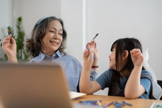 Happy senior grandma and cute little kid granddaughter watching cartoons on laptop together, happy older grandmother grawing and painting