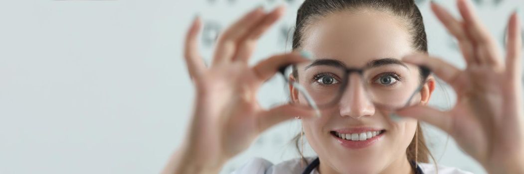 Portrait of woman eye doctor with eye test chart on background. Female optician try on glasses, appointment in eye office. Checkup, ophthalmologist concept