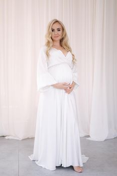 Beautiful pregnant blond hair woman in elegant white dress posing to photographer in studio. Background of white tulle.