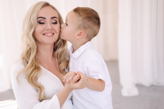 Beautiful pregnant woman with her son. Lady in elegant white dress posing to photographer in studio. Background of white tulle.