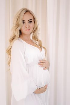 Beautiful pregnant blond hair woman in elegant white dress posing to photographer in studio. Background of white tulle.