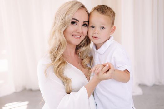 Beautiful pregnant woman with her son. Lady in elegant white dress posing to photographer in studio. Background of white tulle.