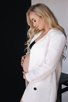 Studio portrait of gogreous pregnant woman in white jacket hold her hand on belly and looks down.