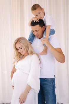 Dad hold his son on neck and hug his pregnat wife. Family look concept for studio photosession. Perents waiting for baby. Happy kid.