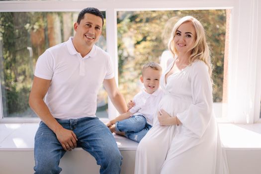 Happy photosession of pregnant woman her husband and their son. Family waiting for baby. White closes family look concept.