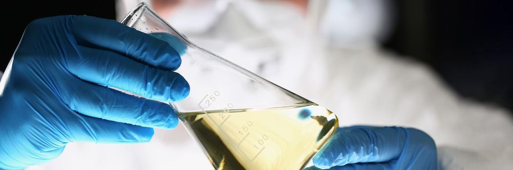 Close-up of laboratory pharmacist in respirator analyzing flask with yellow liquid. Chemist working on test in blue gloves. Chemistry, science, lab concept