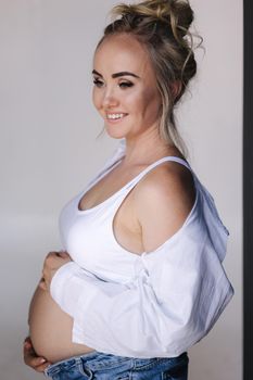 Close up portrait of pregnant woman in studio on white background. Beautiful future mother put her her hands on her belly. Stylish woman in white jaket