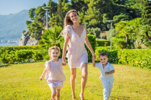 Joyful young mother playing catch together with two little children, boy and girl in green park on a summer day. Childhood, parenting, happiness concept.