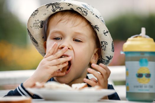 Close up image of Caucasian baby boy with blonde hair and hat opening mouth going to bite crispy bread, having joyful facial expression. Childhood, food, care and health concept
