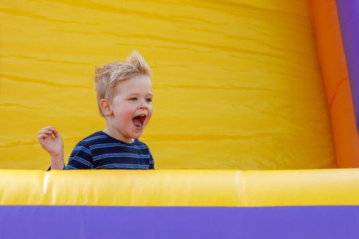 Little boy having fun in inflatable castle playground. Furious child shouts loudly. Bright yellow rubber trampoline background, there is free copy space.