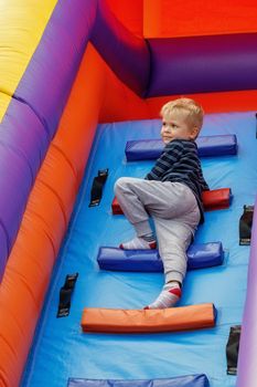 A cute boy climbs a colorful ladder on a high inflatable trampoline and looks into the distance.