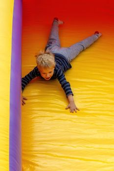 A brave little boy slides down on a red-yellow trampoline at an amusement park.