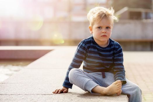 Little sad boy sitting by the fountain on a Sunny day, the child looks to photo camera, there is free space for text in the picture. Summer leisure for kids in the city.