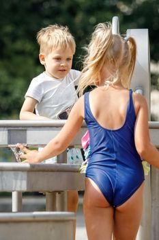 A happy little boy and nice girl in a blue swimsuit plays with a water tap in a city park. Special water equipment for children's games on a hot summer day outdoors. Vertical photo.