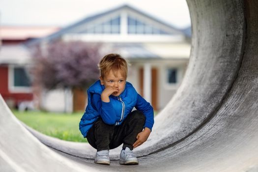 A cute kid wearing a blue vest squats in a concrete tunnel and thinks of something. Horizontal photo.