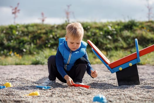 A cute blond child is playing with sand toys near the balance swing in the yard. Safe surface with of small pebbles in playground.