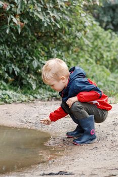 A child in a red jacket and rubber boots explores the mud of a swamp in nature on a hike in the fresh air.