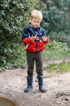 A little boy explores the mud near a puddle during a walk in nature, he looks at his smeared hands.