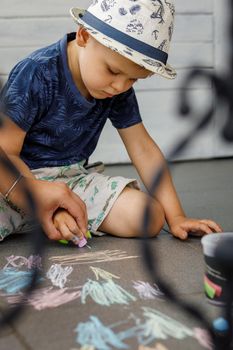 The little boy and his mother draw with colored chalks on the top of a concrete sidewalk. Children's outdoor activities in summer. The mother controls her child's hand.