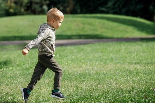 Cute boy running across grass und smiling. An agile happy child is playing sports in a summer city park.