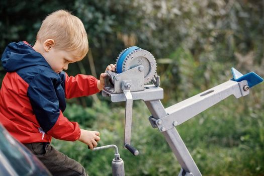 The little boy concentrated and playing with boat trailer winch, boat bow rest, blue cargo strap and its pulley and gears. The child turns the knob.