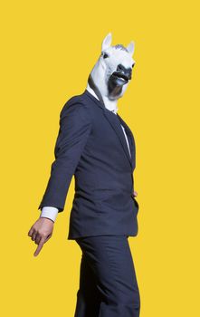 A vertical shot of a creative man in a costume with a white horse mask posing on a yellow background wall.