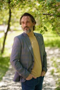 Mature businessman with grey beard wearing casual jacket and jeans, spend time at park or garden looking thoughtful sideways of camera holding hands in pockets.