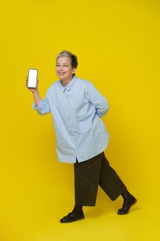 Showing phone in hand mature grey haired woman smiling on camera. Pretty woman in blue shirt and brown trousers isolated on yellow background. Mobile app advertising. Mockup product placement.