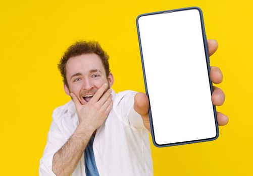 Cant believe, shocked young man with smartphone showing a white empty screen isolated over yellow game, bet, lottery win. Product placement. Mobile App Advertisement.