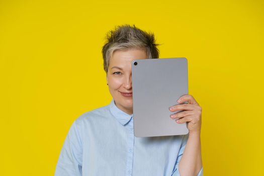 Grey haired woman hide face cover it by digital tablet smiling shy working or shopping online or checking on social media. Pretty woman in blue shirt isolated on yellow background.