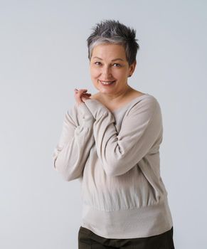 Mature tender grey hair woman in 50s european ethnicity posing looking at camera with hands folded wearing cream blouse isolated on white. Smiling woman healthcare, aging beauty, cosmetics concept.