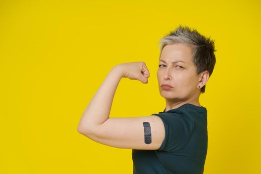 Health is power senior woman with grey hair showing nude bicep, shoulder with band aid or plaster on it isolated on yellow background. Amazing mature woman 50s vaccination and healthcare concept.