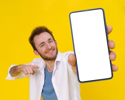 Young man pointing at smartphone showing a white empty screen game, bet, lottery win isolated over yellow background. Product placement for mobile application advertisement.