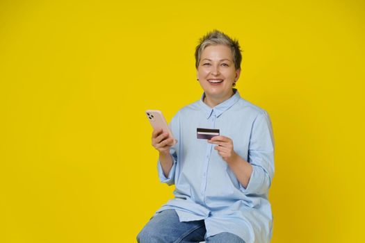 Grey haired woman hold phone and debit or credit card in hand making online payment or shopping. Pretty mature woman in blue shirt and jeans isolated on yellow background.