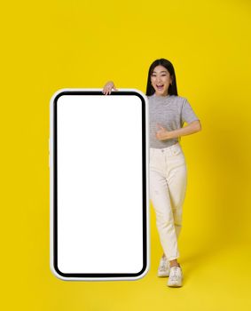Charming asian girl gesturing thumb up stand near huge, giant smartphone with white screen happy dressed in casual isolated on yellow background. Free space mock up. Product placement mobile app add.
