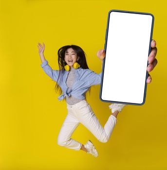 Happy jumping asian girl with smartphone in hand showing white screen mobile app advertisement isolated on yellow background. Full length portrait of joyful asian girl. Product placement.