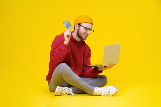 Young man sit on the floor holding laptop and debit or credit bank card wearing red hoodie and yellow hat buying or paying online. Smart freelancer man with laptop isolated on yellow background.